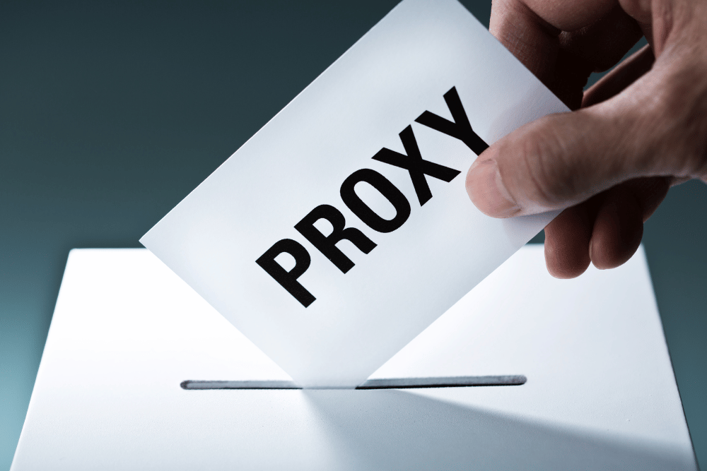 A human hand placing a white card with the words 'Proxy' into the slot of a ballot box.