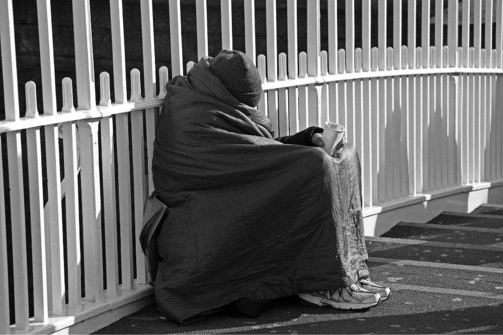 Outdoor black and white image of a person sitting on steps wrapped in a blanket and wearing a woolly hat